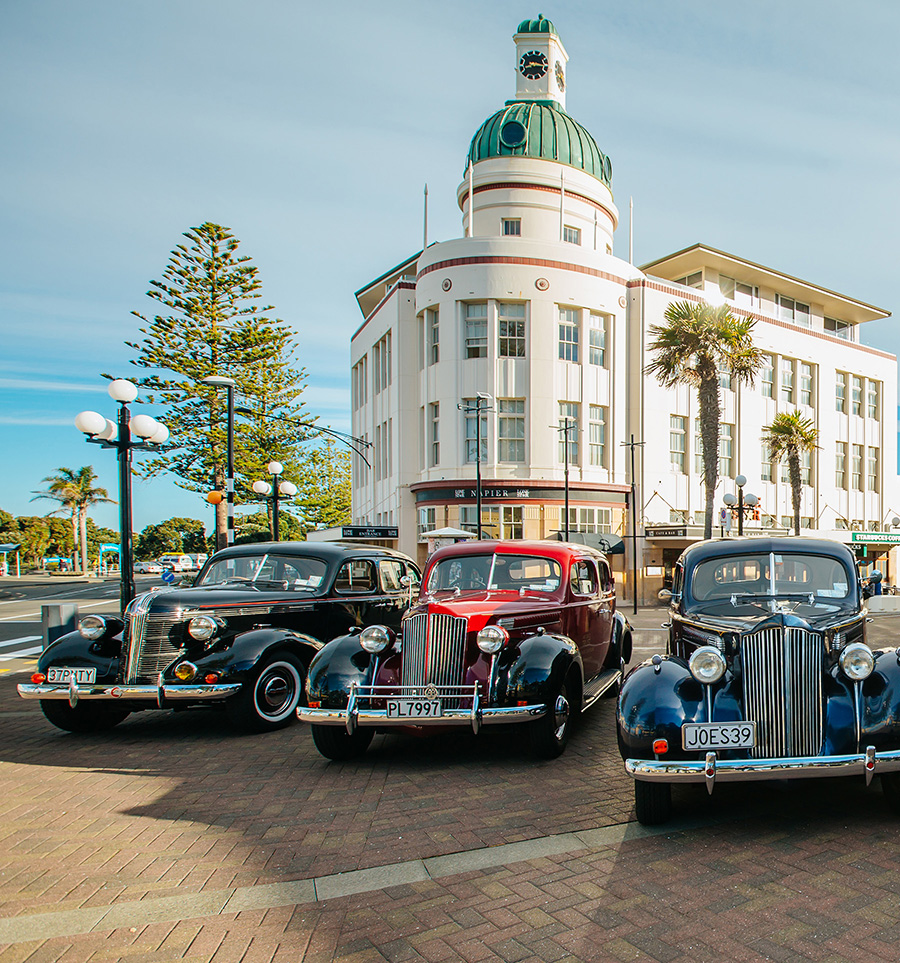 Three vintage cars are parked in front of an art deco building