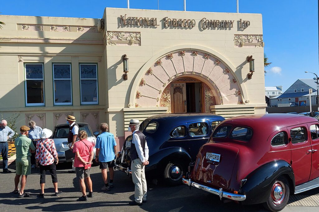 A group of tourists look at an art deco building with vintage cars parked to the side