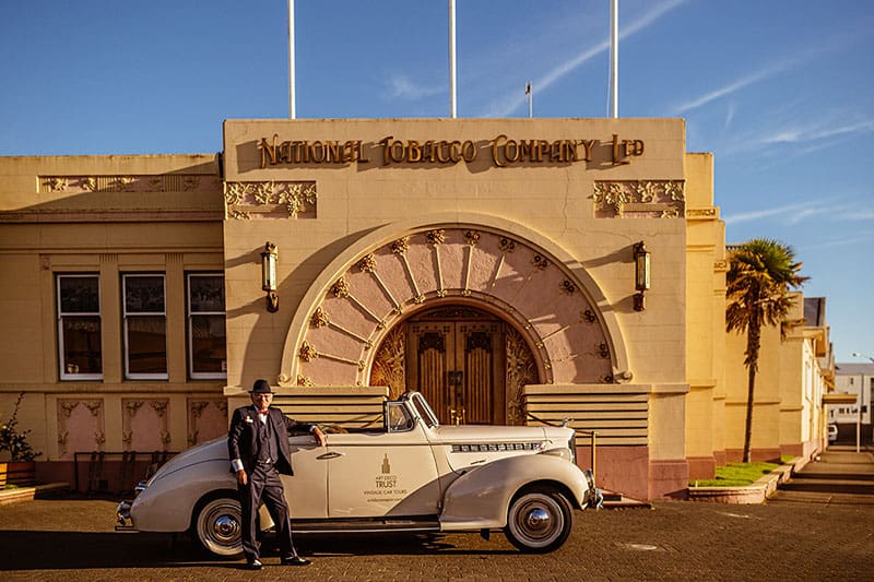 A man poses in front of a cream colored vintage car in front of an art deco building